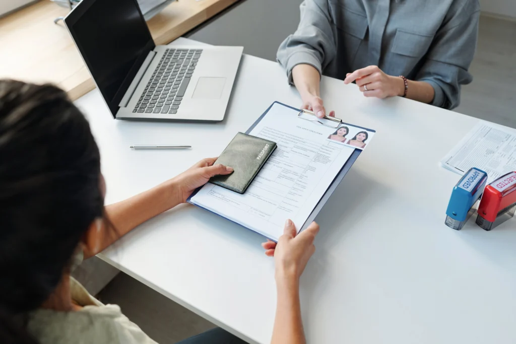 A person hands over a visa application form and a passport to another individual at a desk with a laptop and stamping tools, representing the Document Attestation process.